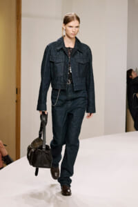 Model walking a runway in a dark denim jean set: jacket, high-waisted jeans, and brown leather bag in hand, brown shoes visible.