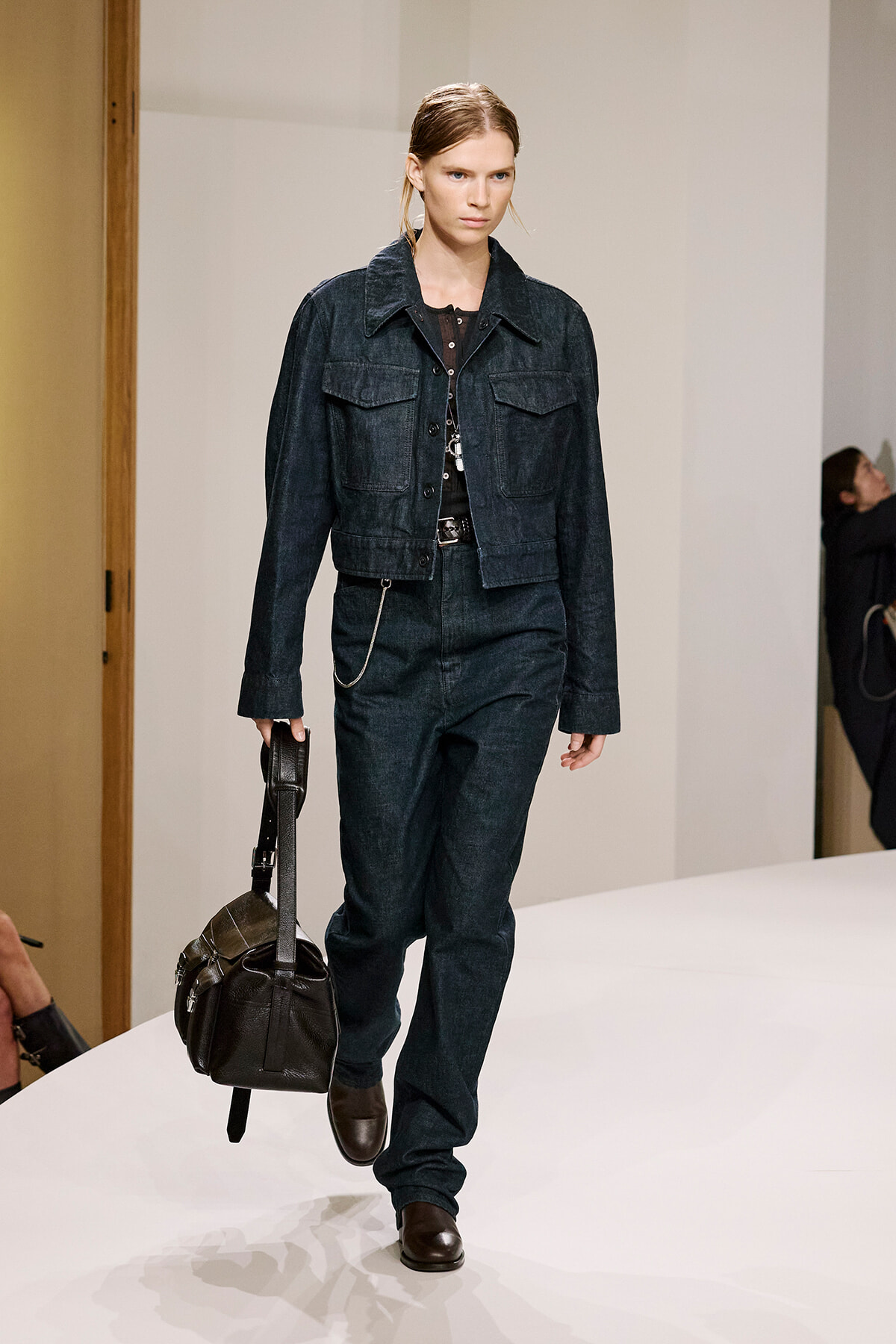 Model walking a runway in a dark denim jean set: jacket, high-waisted jeans, and brown leather bag in hand, brown shoes visible.