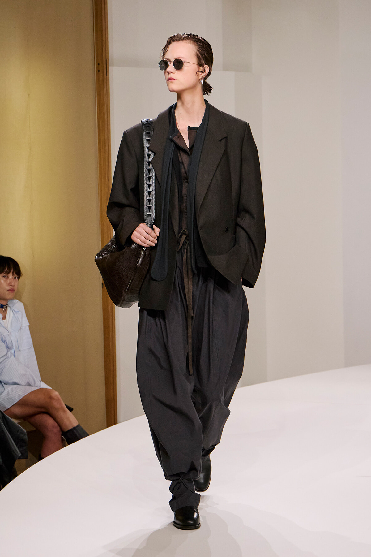 Model walking the runway in an oversized black suit, sheer top, black pants, and sunglasses, carrying a large black bag with a studded strap.