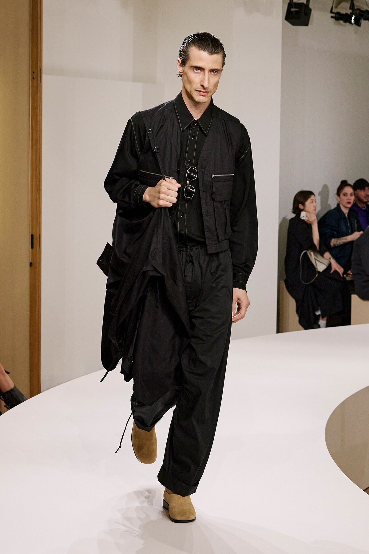 Male model in black utility outfit walking a white runway, holding a black coat and sunglasses hanging from his shirt, tan boots visible.