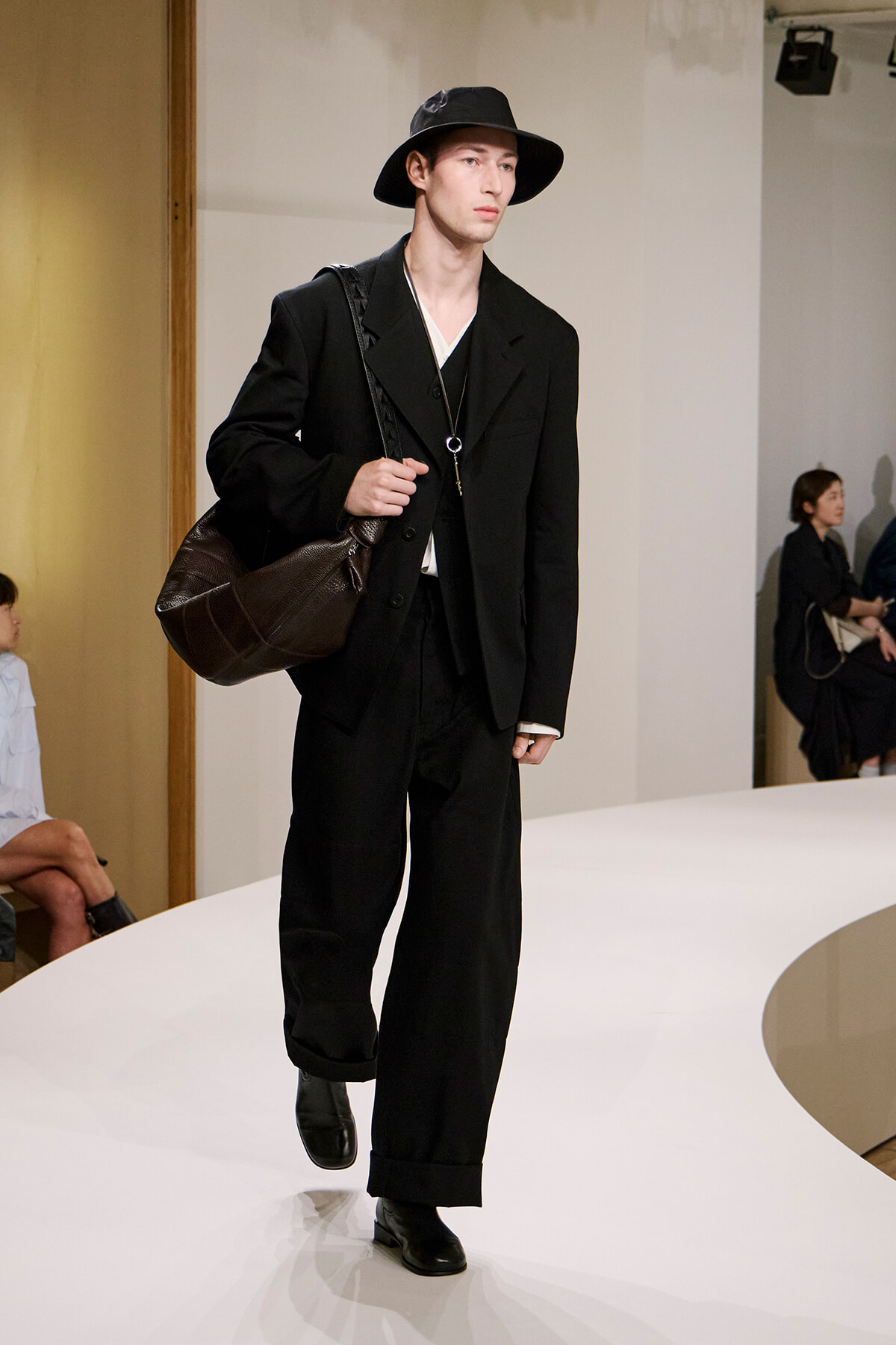 Male model walking a fashion runway in a black suit with a wide-brim hat, carrying a large brown shoulder bag.