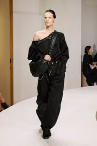 Model walking a runway in an oversized black one-shoulder jumpsuit with a diagonal neckline and a large black crossbody bag