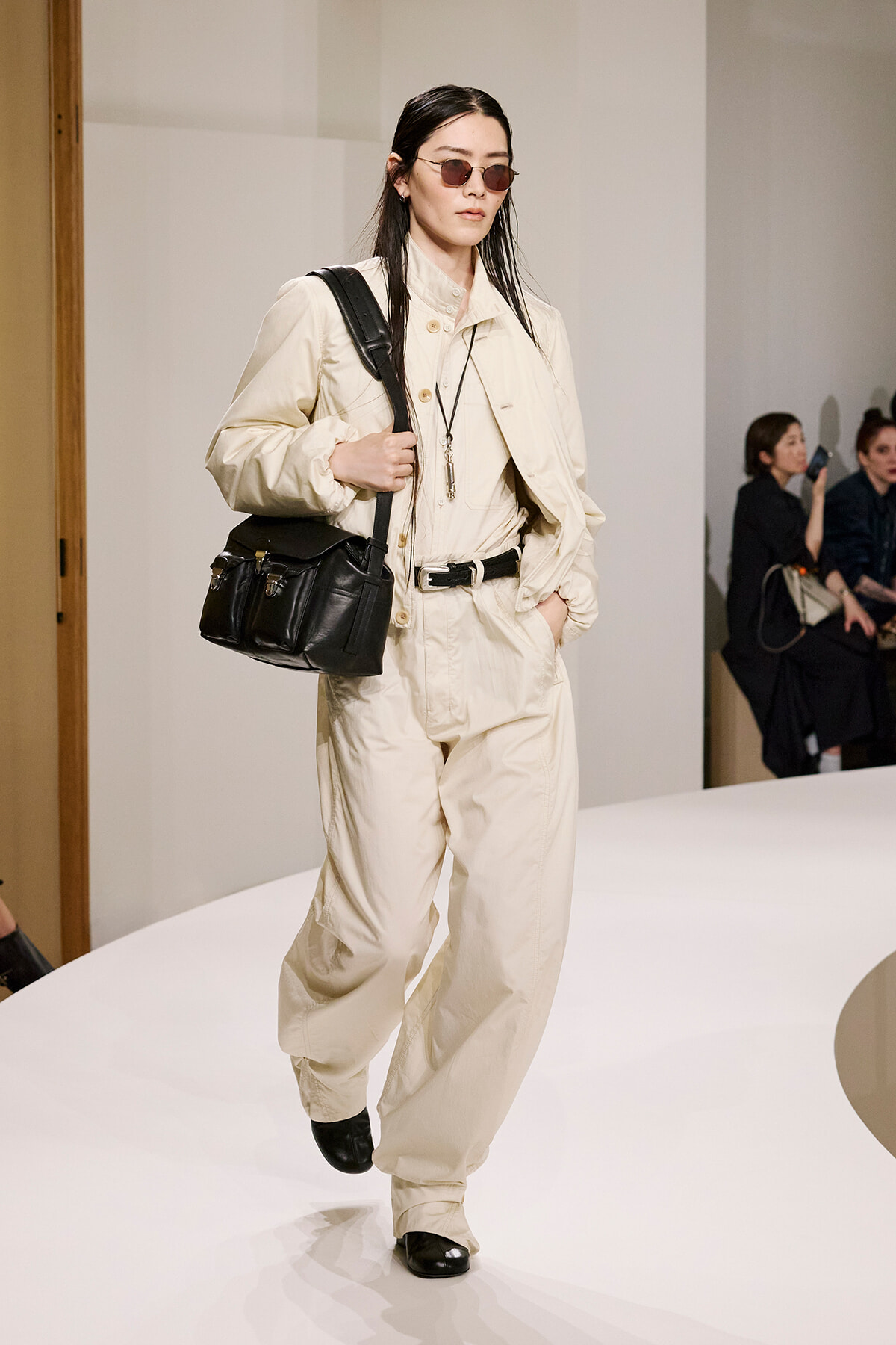 Model walks the runway in a cream-colored jumpsuit with a black shoulder bag and tinted sunglasses.