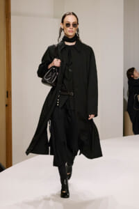 Model walking a white runway in an all-black outfit: long coat, layered black pieces, black boots, and a black handbag.