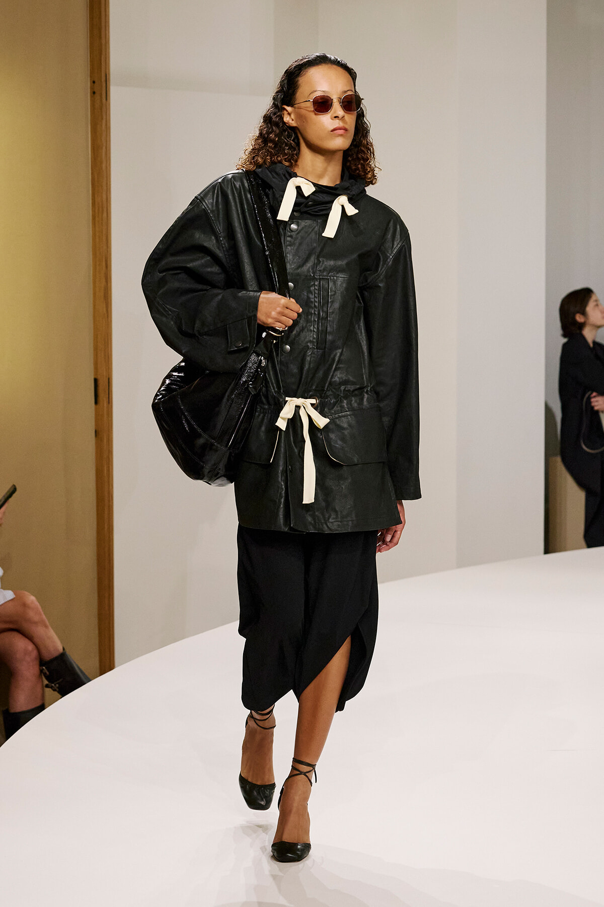 Model walking a runway in a oversized black leather jacket with cream drawstrings, black skirt, and strappy heels, wearing round sunglasses.