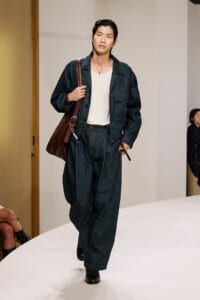 Male model wearing a dark denim suit with a white shirt, carrying a brown shoulder bag on a runway.