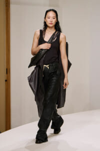 Model walking a runway in an all-black outfit: sleeveless top, loose black pants, and a long draped coat, with a dark crossbody bag over the shoulder.
