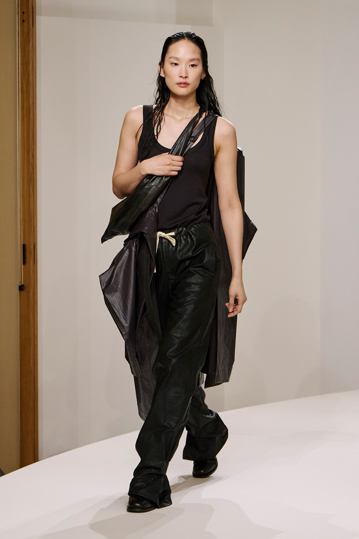 Model walking a runway in an all-black outfit: sleeveless top, loose black pants, and a long draped coat, with a dark crossbody bag over the shoulder.