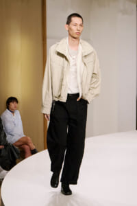 Model walking a runway in a beige oversized jacket, white shirt, and black wide-leg pants with black boots.