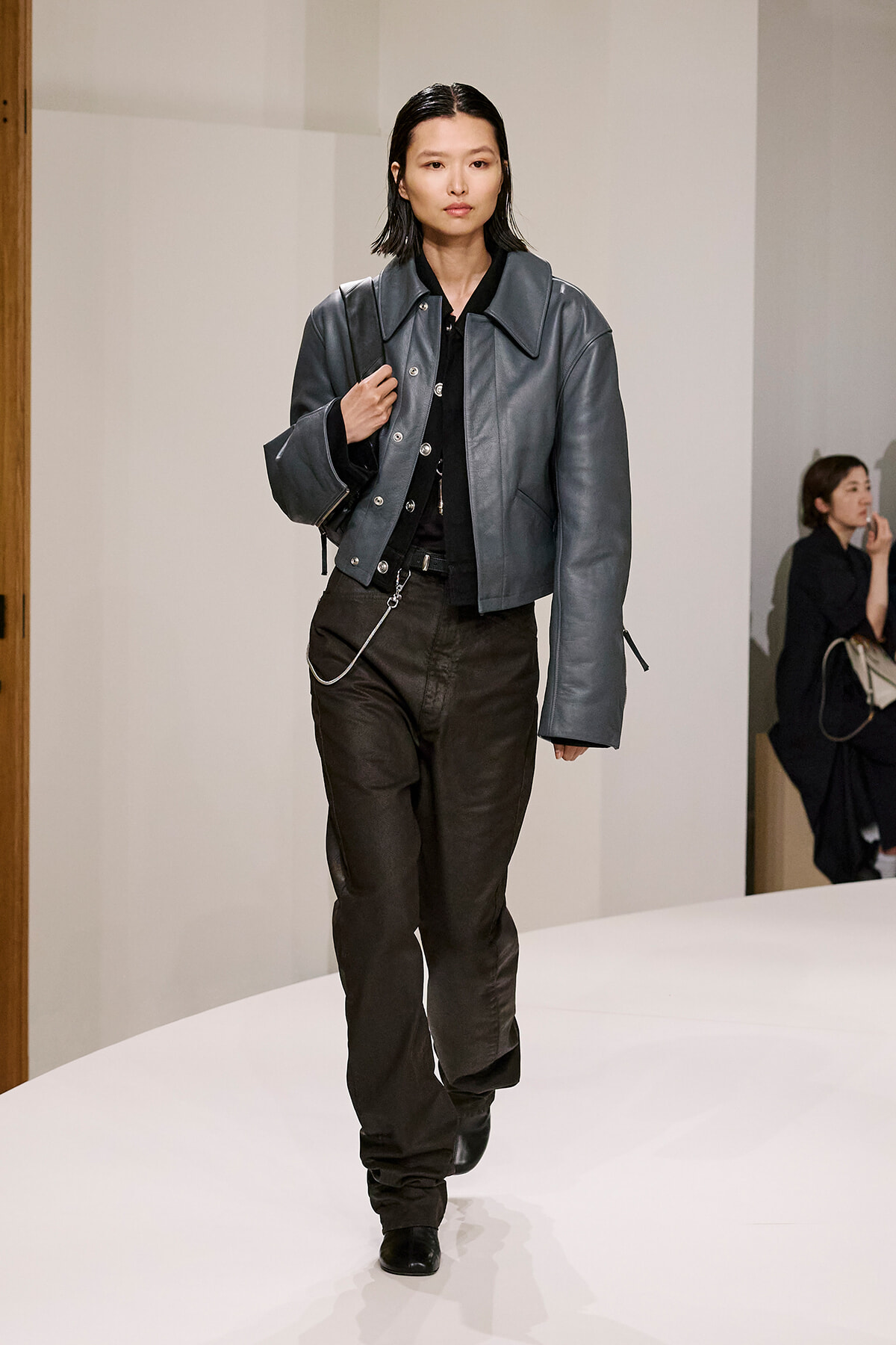 Model walking a runway in a gray leather cropped jacket over black top and dark cargo-style pants; minimalist backstage setting.