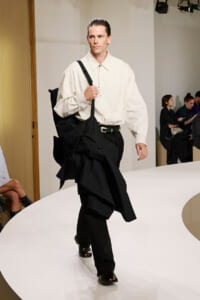 Male model walking a runway in a cream shirt and black trousers, carrying a black coat-slash-bag over his shoulder on the catwalk.
