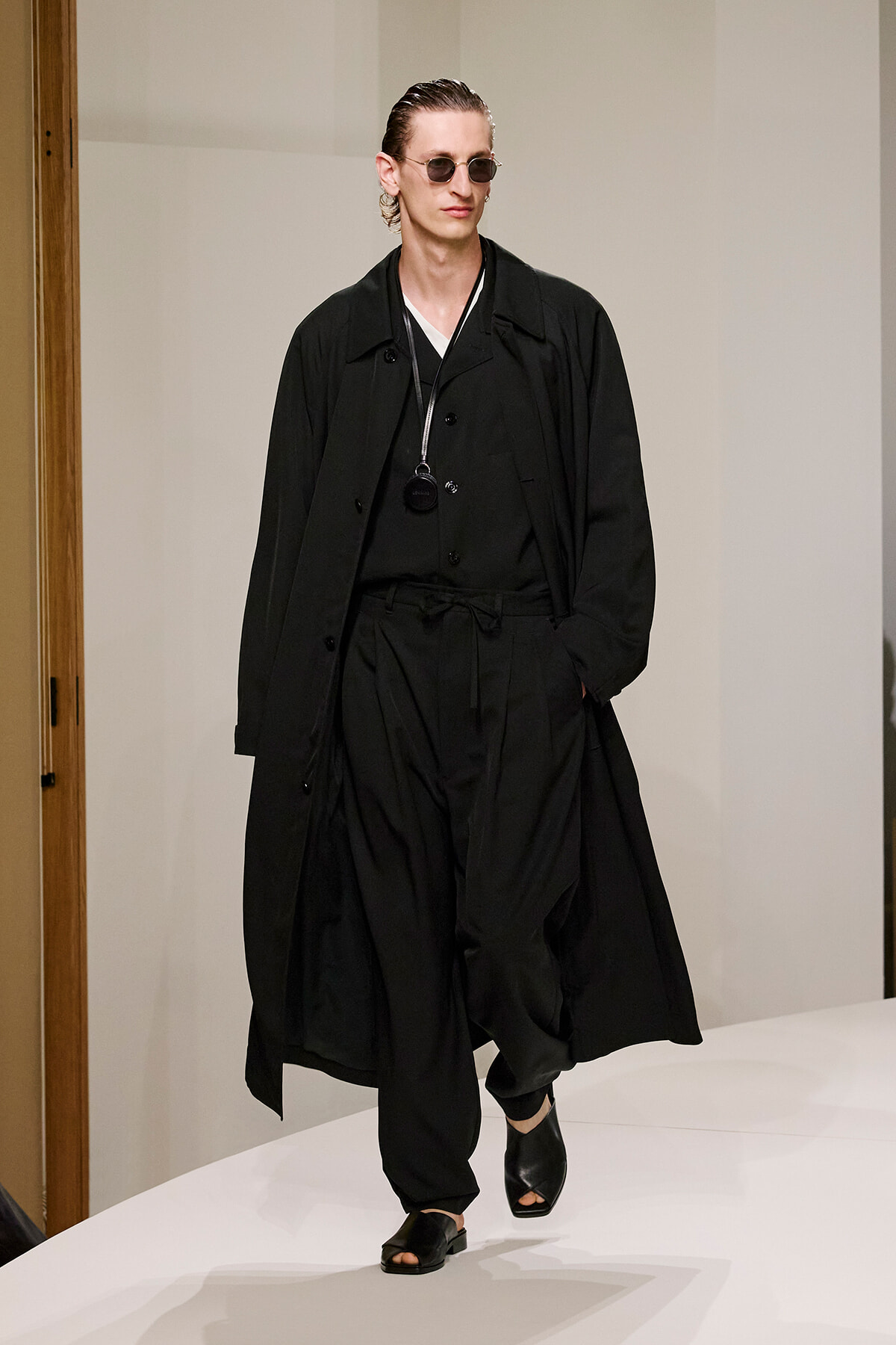 Male model in a full black ensemble (trench coat, layered top and pants) wearing sunglasses and a pendant, walking a runway.