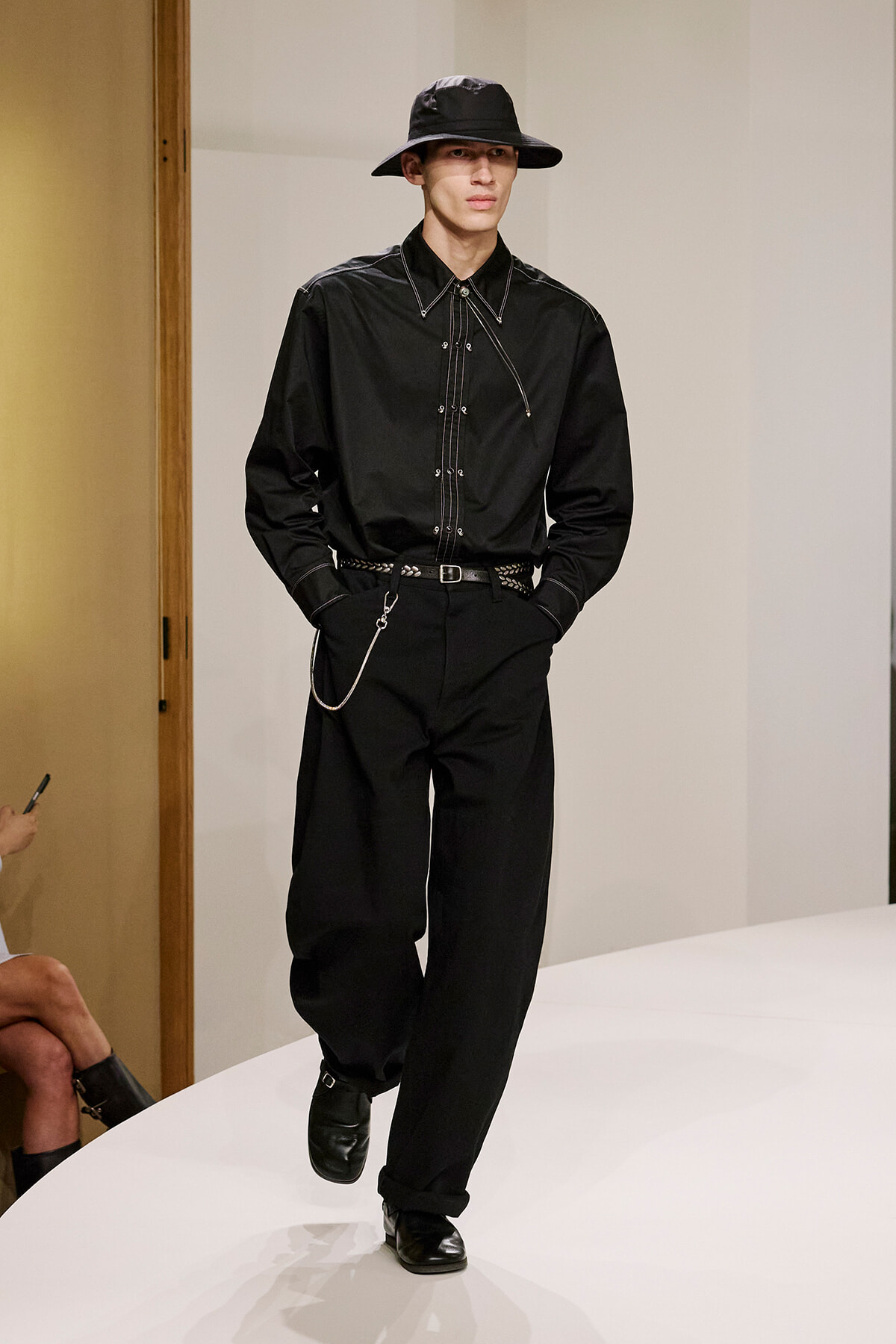Male model on a runway in an all-black Western-inspired outfit: wide-brim hat, button-down shirt with white piping, belt with a chain, and black pants.