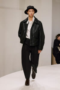 Male model walks a white runway in a black leather jacket, white shirt, black trousers, and a black hat at a fashion show.