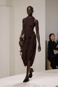Model walking a runway in a dark burgundy sleeveless dress with beaded harness and matching accessories, carrying a woven bag.