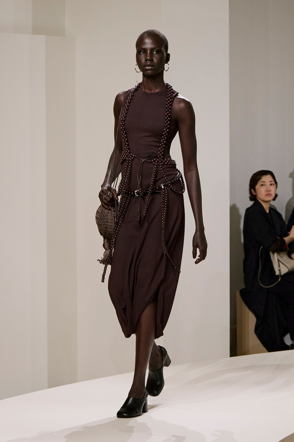 Model walking a runway in a dark burgundy sleeveless dress with beaded harness and matching accessories, carrying a woven bag.