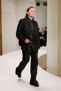 Model walking a white runway in an all-black outfit with a crossbody bag and studded belt.