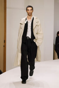 Model walking runway in a long beige trench over a white shirt, black vest and wide black trousers, black shoes.