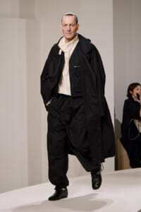 Male model walking a runway in an oversized black coat, white top, and loose black pants with black boots, looking serious.