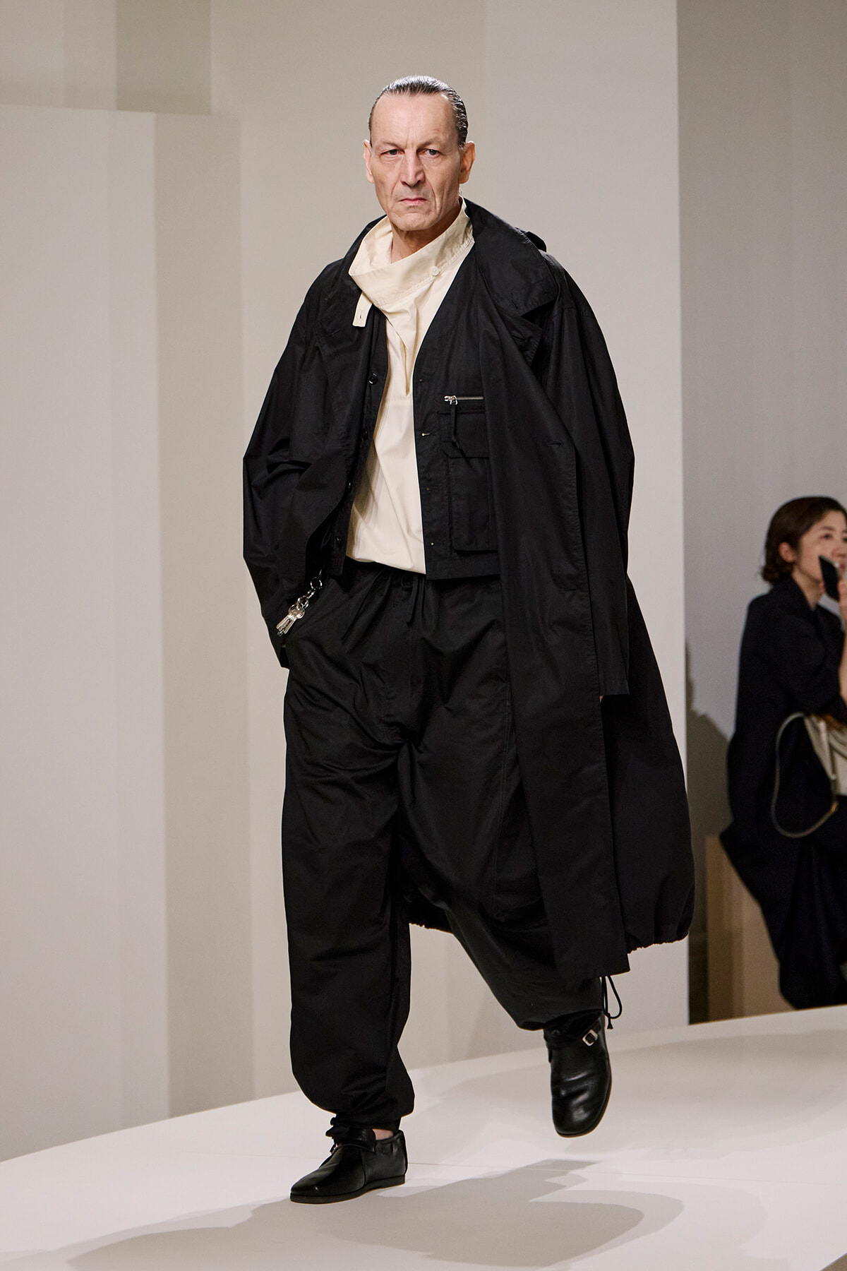 Male model walking a runway in an oversized black coat, white top, and loose black pants with black boots, looking serious.