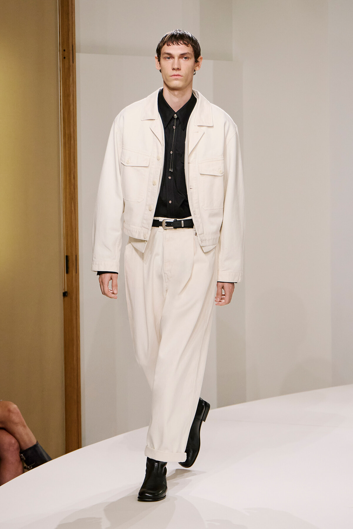 Male model walking a fashion show in a cream jacket and matching wide-leg pants with a black shirt and belt on a runway.