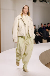 Model walking a white fashion runway in a cream oversized jacket and light green baggy pants.