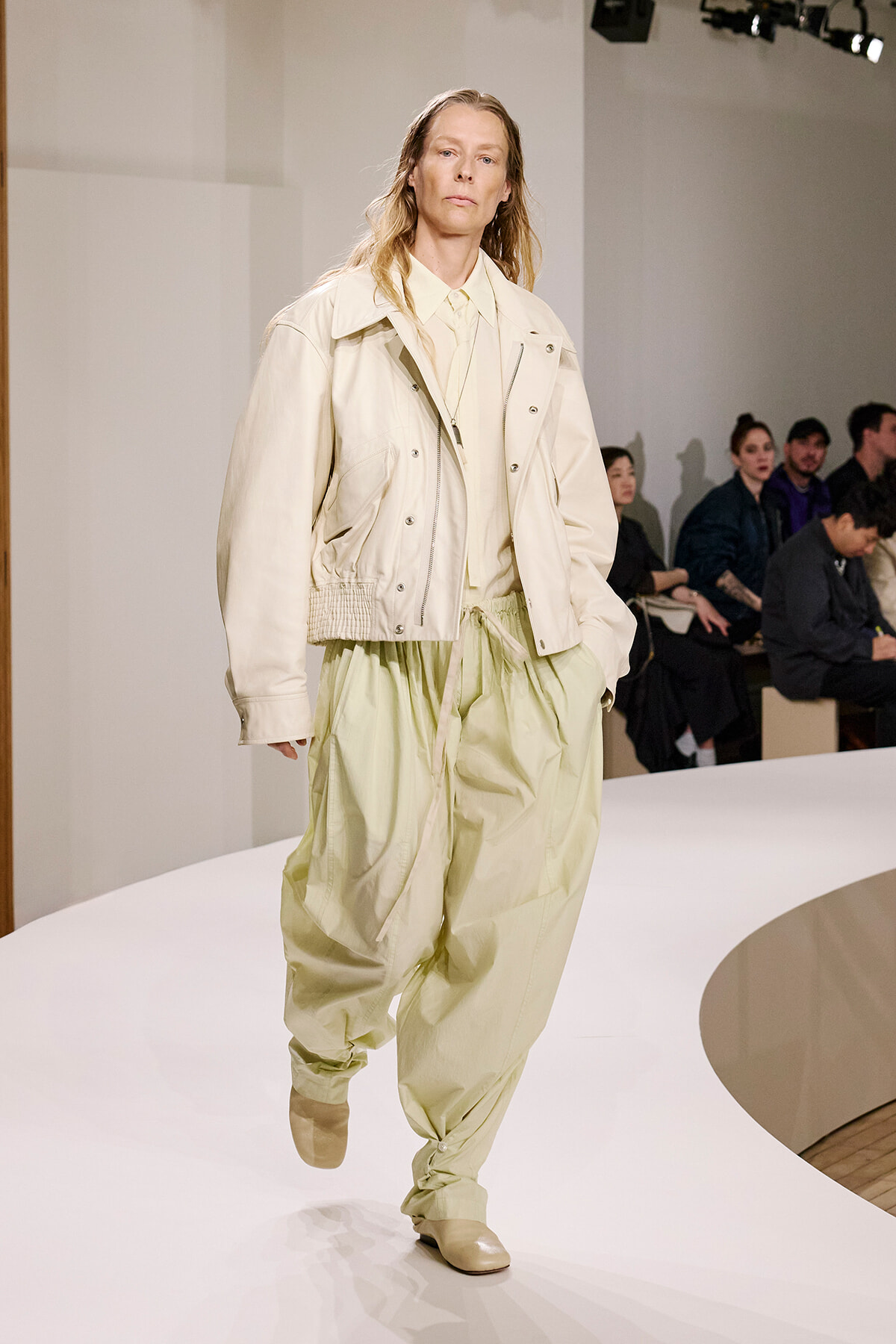 Model walking a white fashion runway in a cream oversized jacket and light green baggy pants.