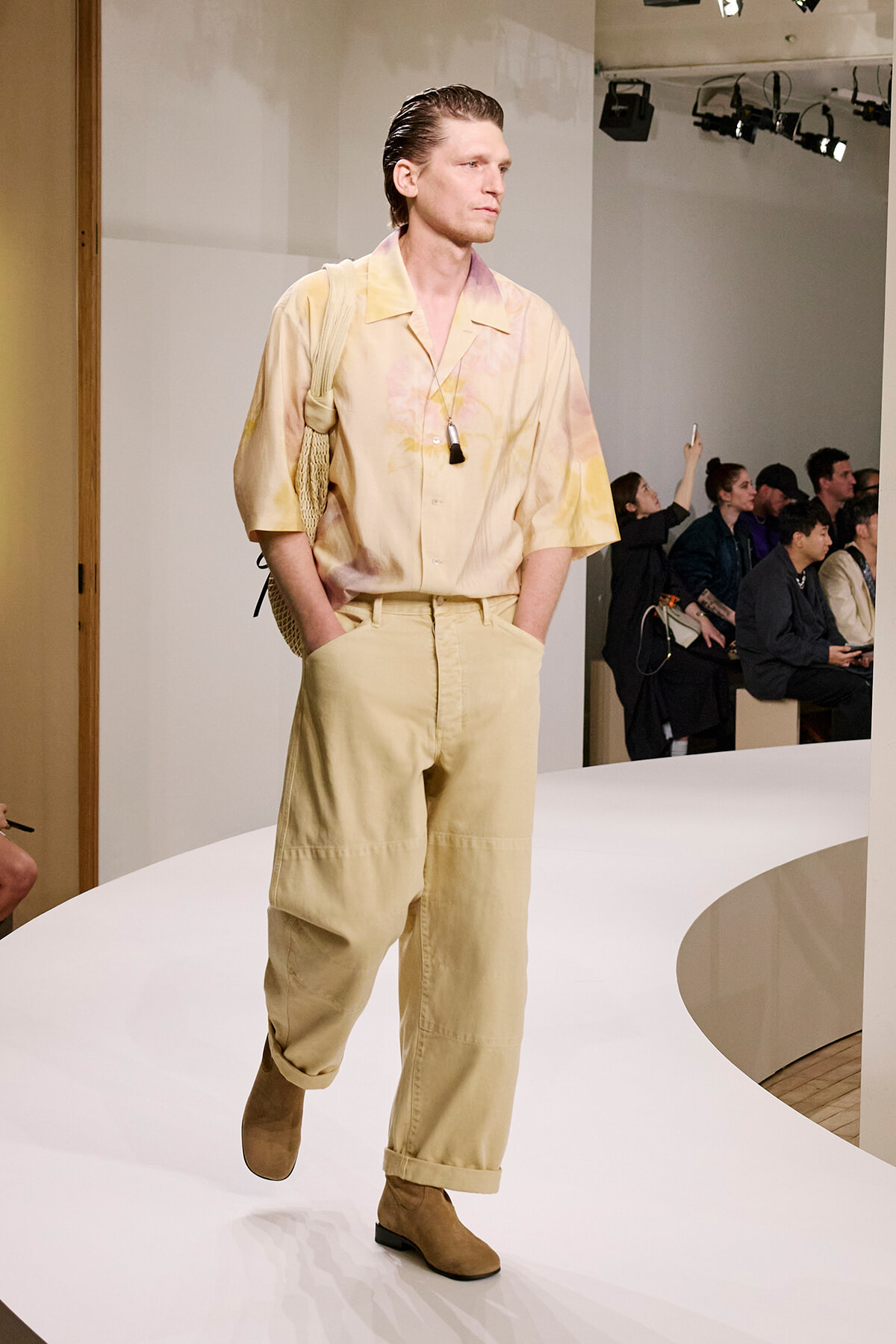 Male model walks a fashion runway in a beige, tie-dyed shirt with rolled sleeves, matching wide-legged pants, tan boots, and a woven bag over the shoulder; audience in background.