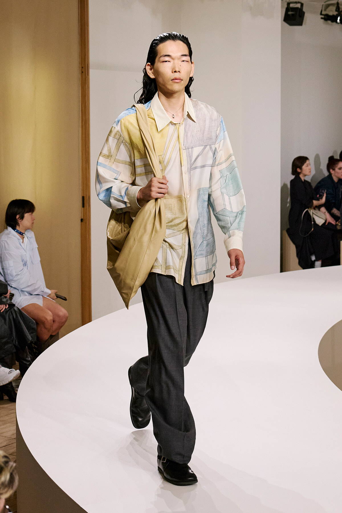 Male model walks a white runway wearing a pastel patchwork shirt, beige tote over one shoulder, and dark trousers.