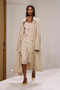 Model walks a beige-toned runway in a matching trench coat over a relaxed midi dress, accessorized with a pendant necklace and wraparound heels.