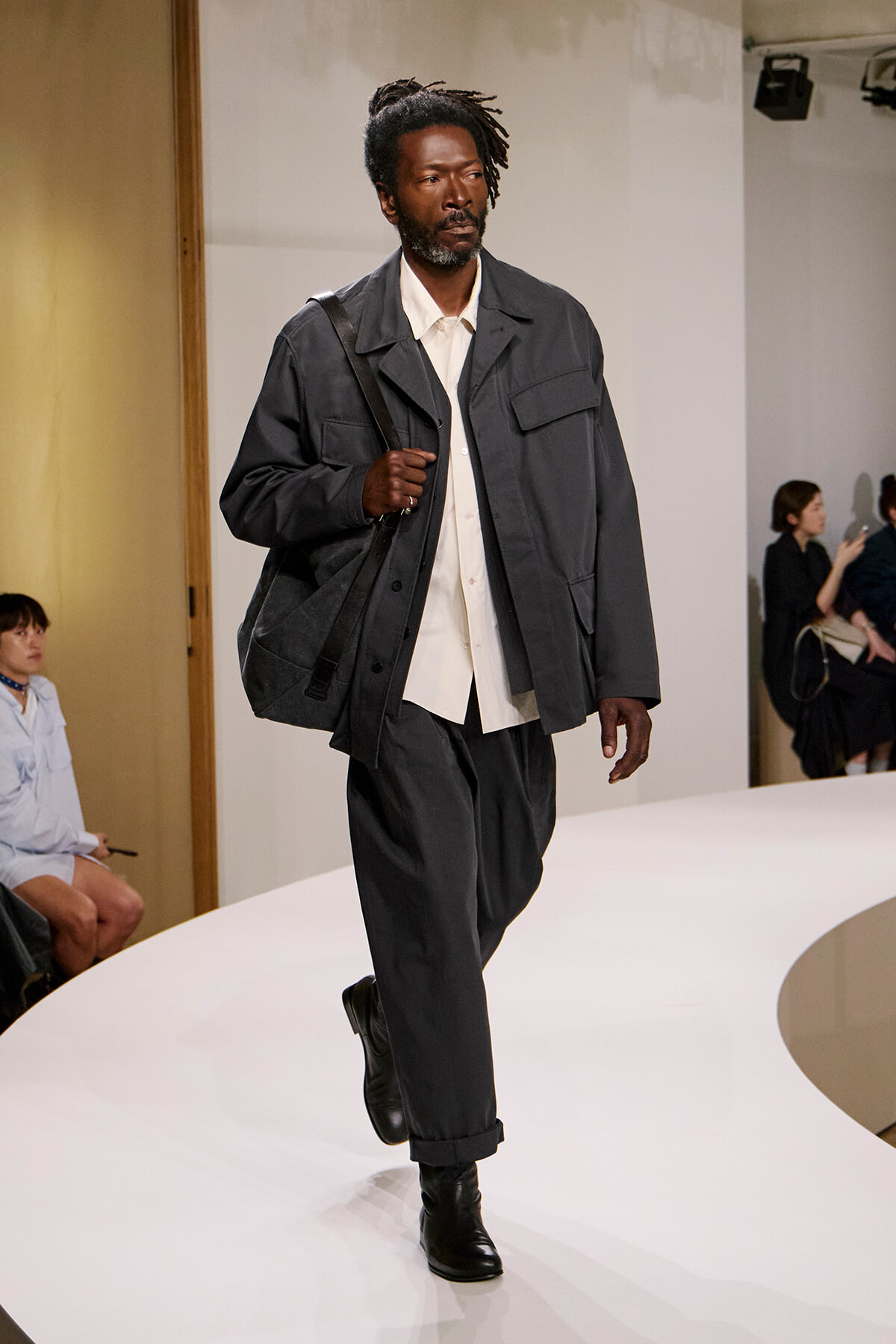 Male model walking a fashion runway, carrying a black shoulder bag, wearing a dark gray utility jacket, cream shirt, and black pants with boots.