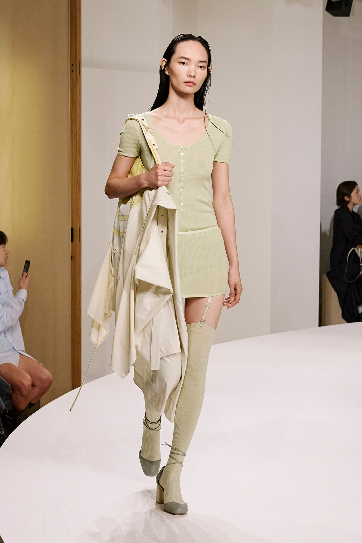 Model walks a runway in a mint-green knit two-piece set, holding a light-colored coat over her arm with matching knee-high socks and heeled shoes.