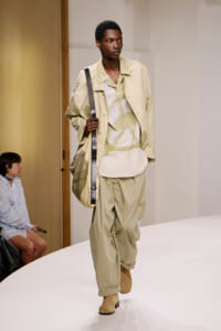 Black model on a runway wearing a beige oversized coat over a pale geometric top and baggy pants, carrying a large black-strapped bag across the body.