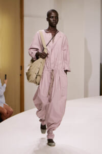 Model walking a fashion show in a pale pink, oversized jumpsuit with a gathered waist, paired with a large beige shoulder bag.