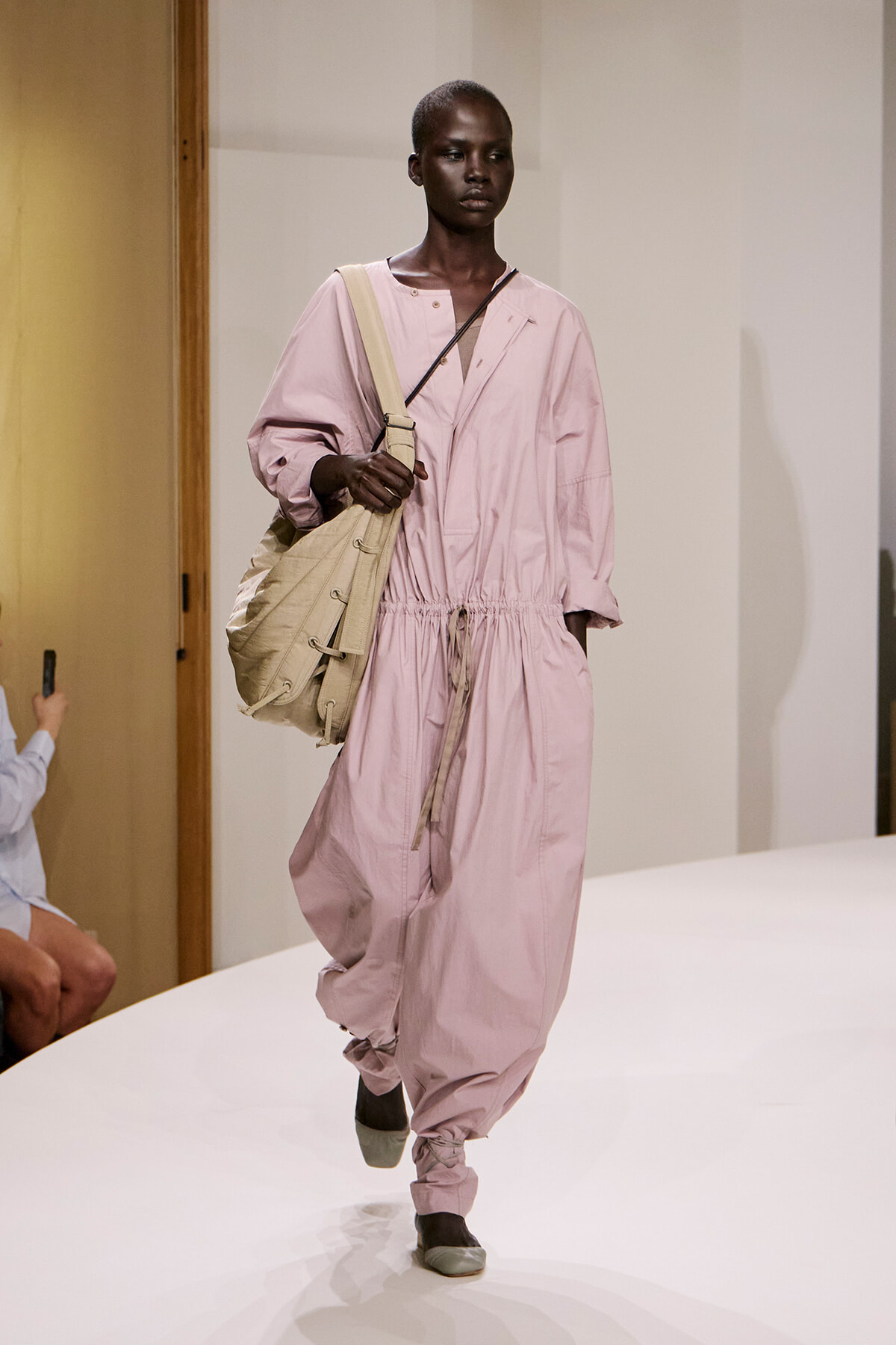 Model walking a fashion show in a pale pink, oversized jumpsuit with a gathered waist, paired with a large beige shoulder bag.