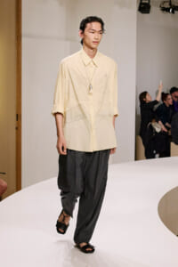 Male model on a white runway wearing a pale yellow oversized shirt with geometric stitching and dark wide trousers; black sandals, pendant necklace visible amid an audience in the background.