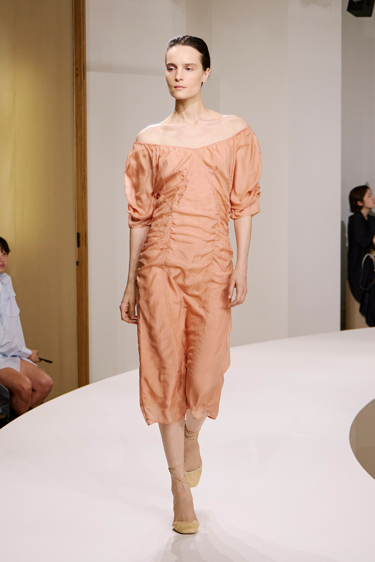 Model walks the runway in a peach satin off-the-shoulder midi dress with ruched sides and beige ankle-strap heels.