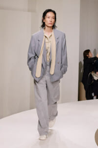 Model in an oversized light-gray suit with a cream scarf on a white runway, neutral expression.