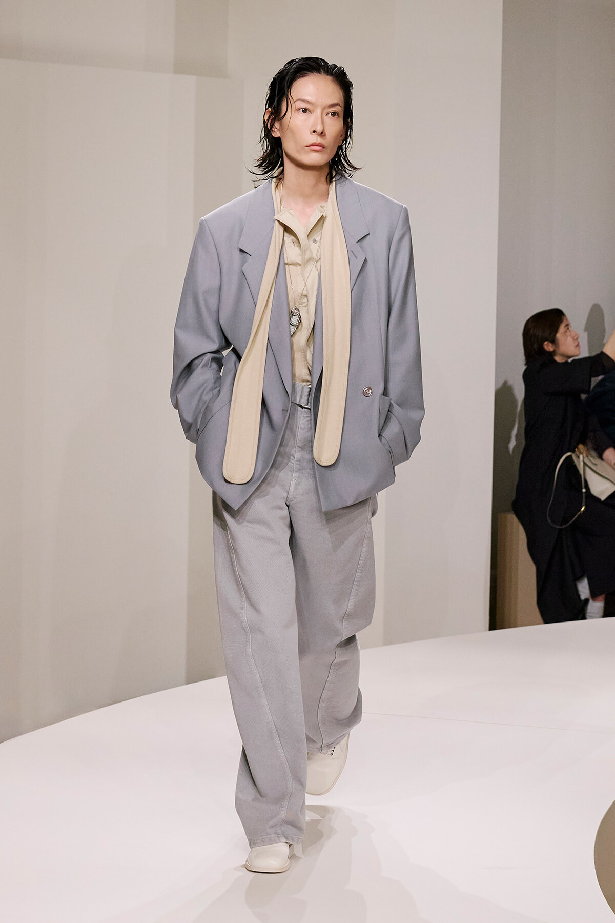 Model in an oversized light-gray suit with a cream scarf on a white runway, neutral expression.