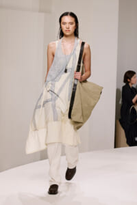 Model on a runway wearing a sleeveless cream dress with pale blue panel details, carrying a large beige tote with a black strap.