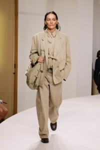Model walking a beige coordinated suit with a large beige bag on a white runway, mid-stride.