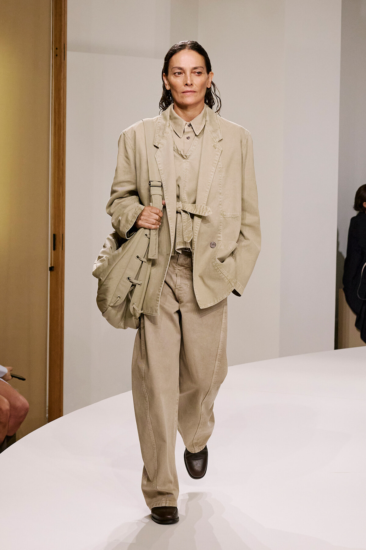 Model walking a beige coordinated suit with a large beige bag on a white runway, mid-stride.