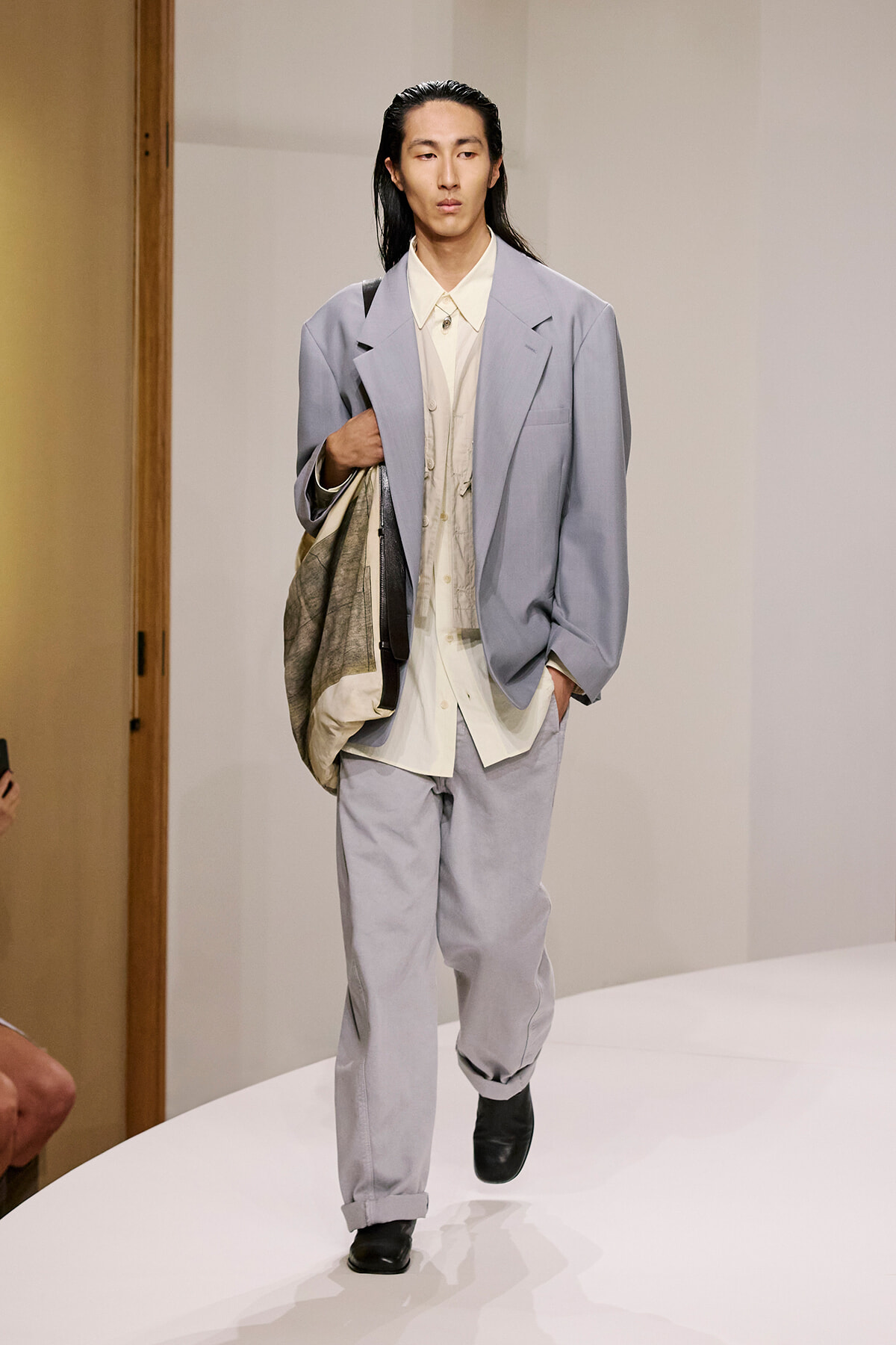 Male model walks runway in an oversized light gray suit with cream shirt, carrying a large bag on the shoulder.