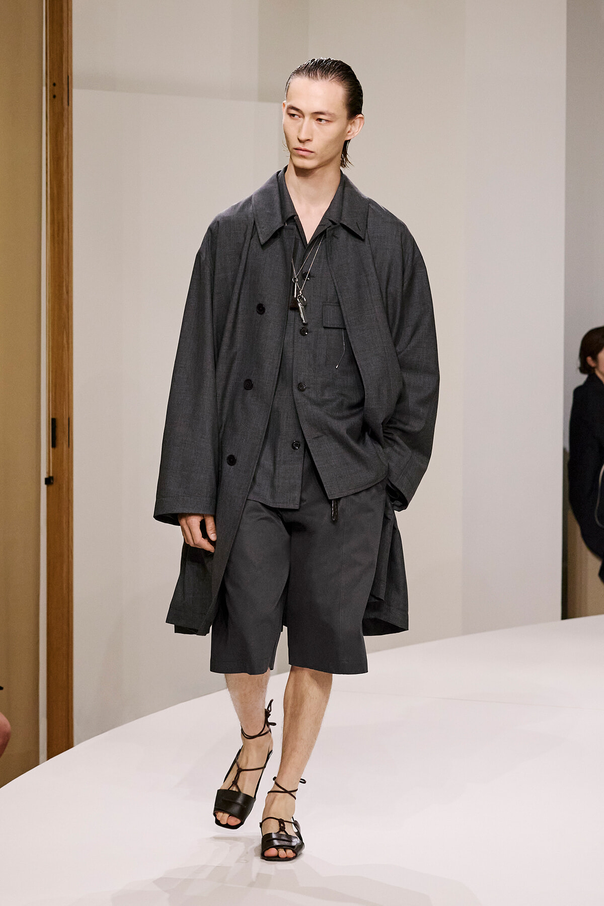 Male model walking a white runway in a dark gray layered outfit: long coat, shirt, and knee-length shorts with black strappy sandals.