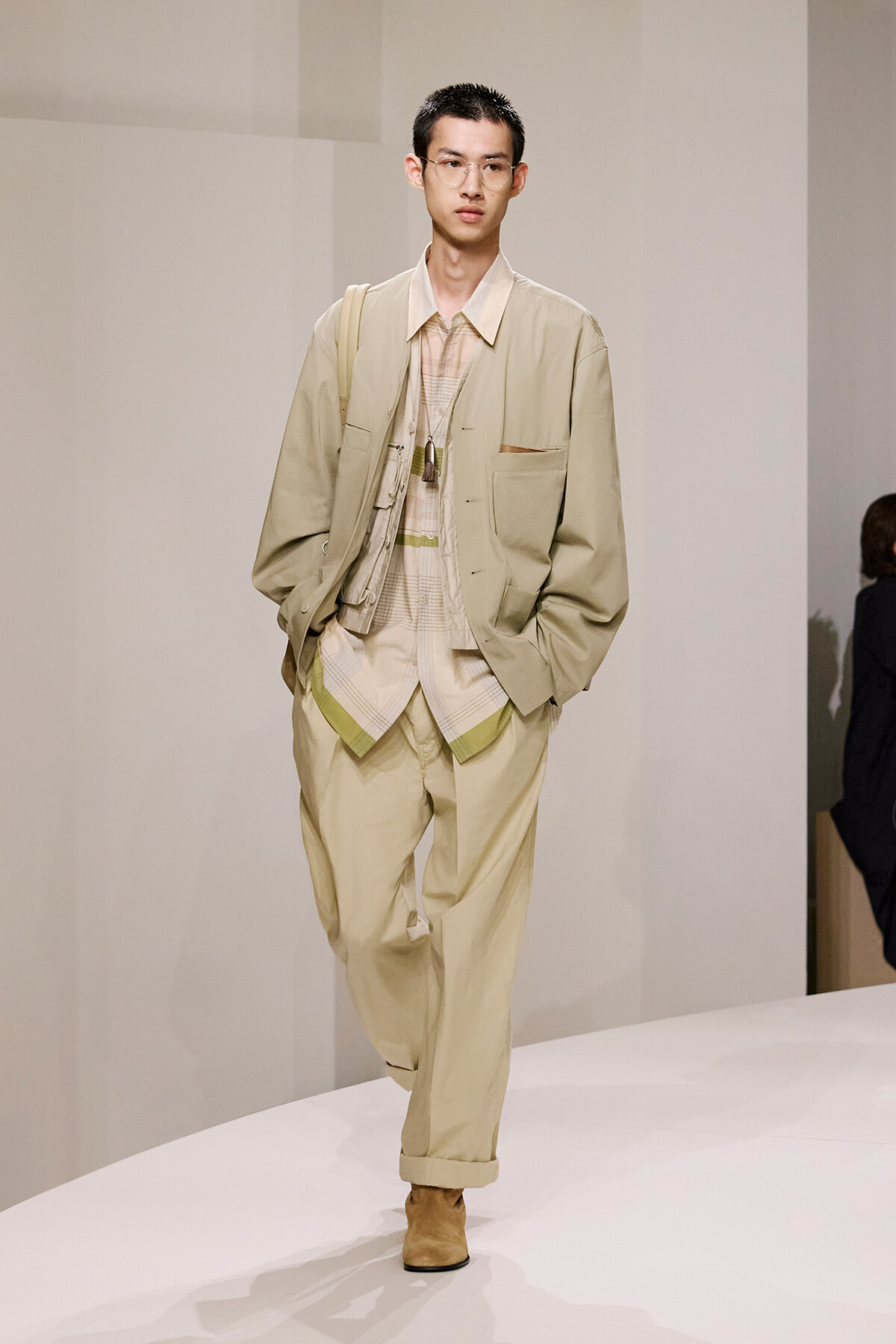 Male model on a white runway wearing a beige layered outfit: oversized jacket, plaid shirt, matching pants, tan boots, and a shoulder bag.