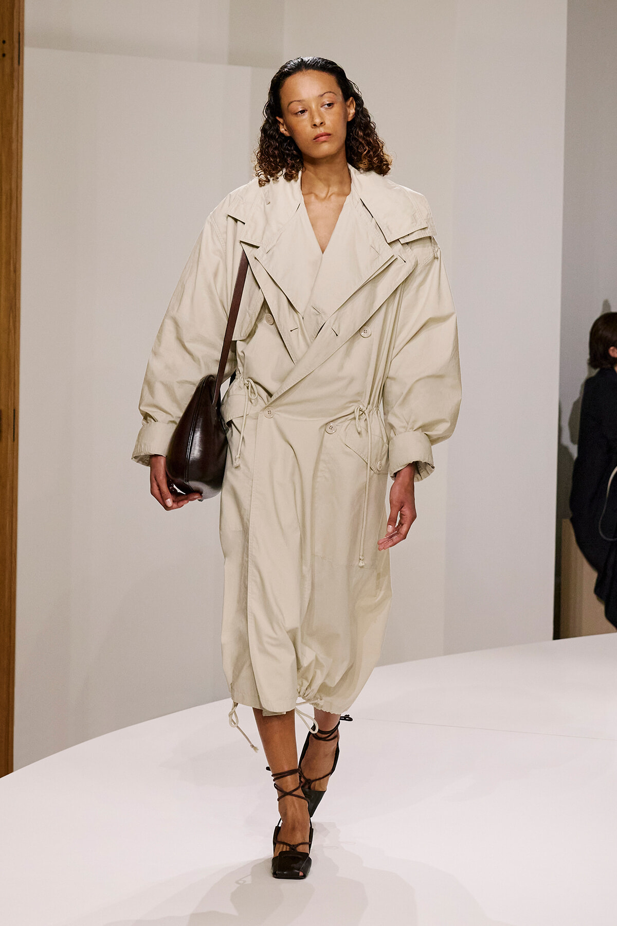 Model walks the runway in an oversized beige trench coat, with a dark brown shoulder bag and black strappy heels.