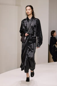 Model walks a black satin wrap dress with a crossbody bag and black mules on a minimalist runway.