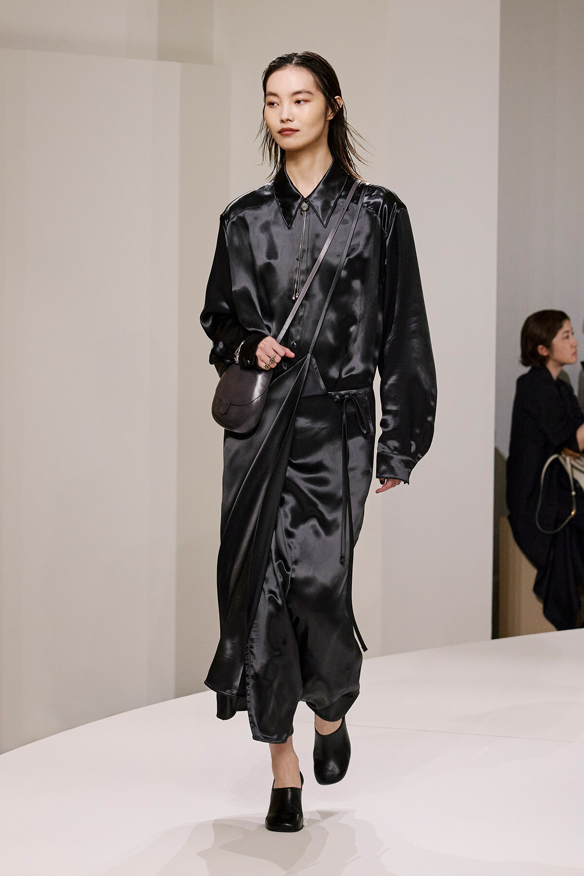 Model walks a black satin wrap dress with a crossbody bag and black mules on a minimalist runway.