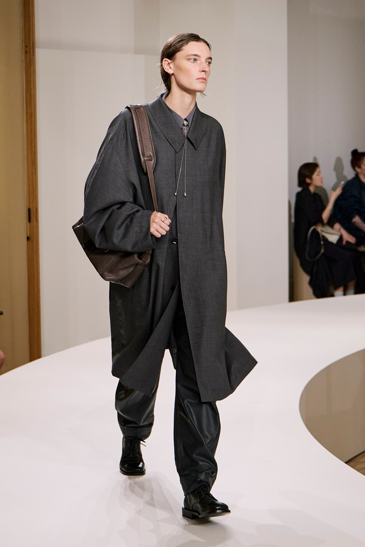 Model walking a runway in a dark gray oversized coat, black pants, and black boots, carrying a brown backpack