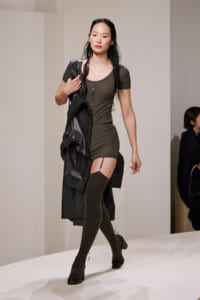 Model walking the runway in a fitted olive-brown mini dress, black thigh-high socks, black heels, and a black coat draped over the shoulder.